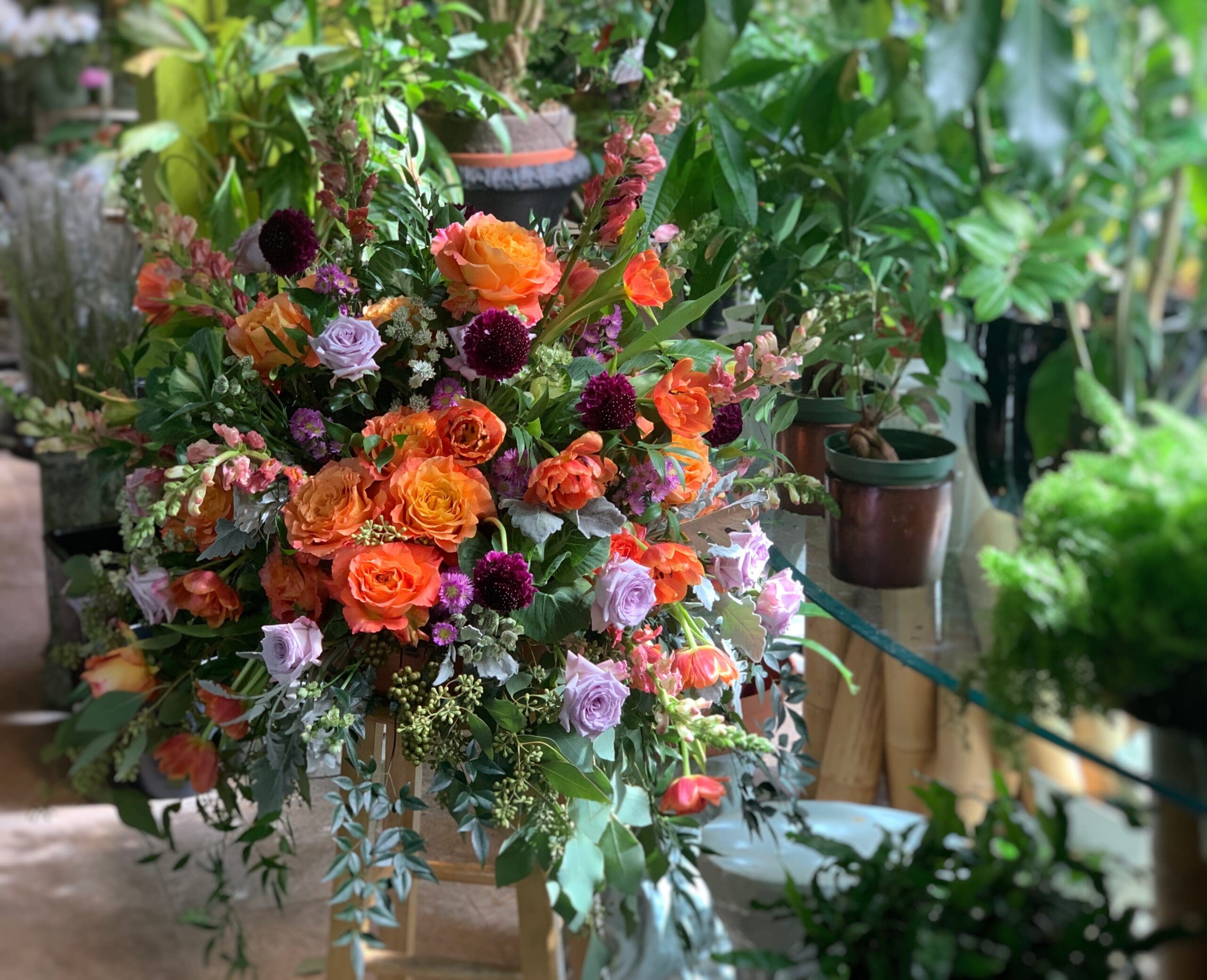 Bold Floral Bouquet with Jewel Tone Pairings Bold floral arrangement with orange, burgundy, and lavender flowers in a terra cotta pot surrounded by greenery
