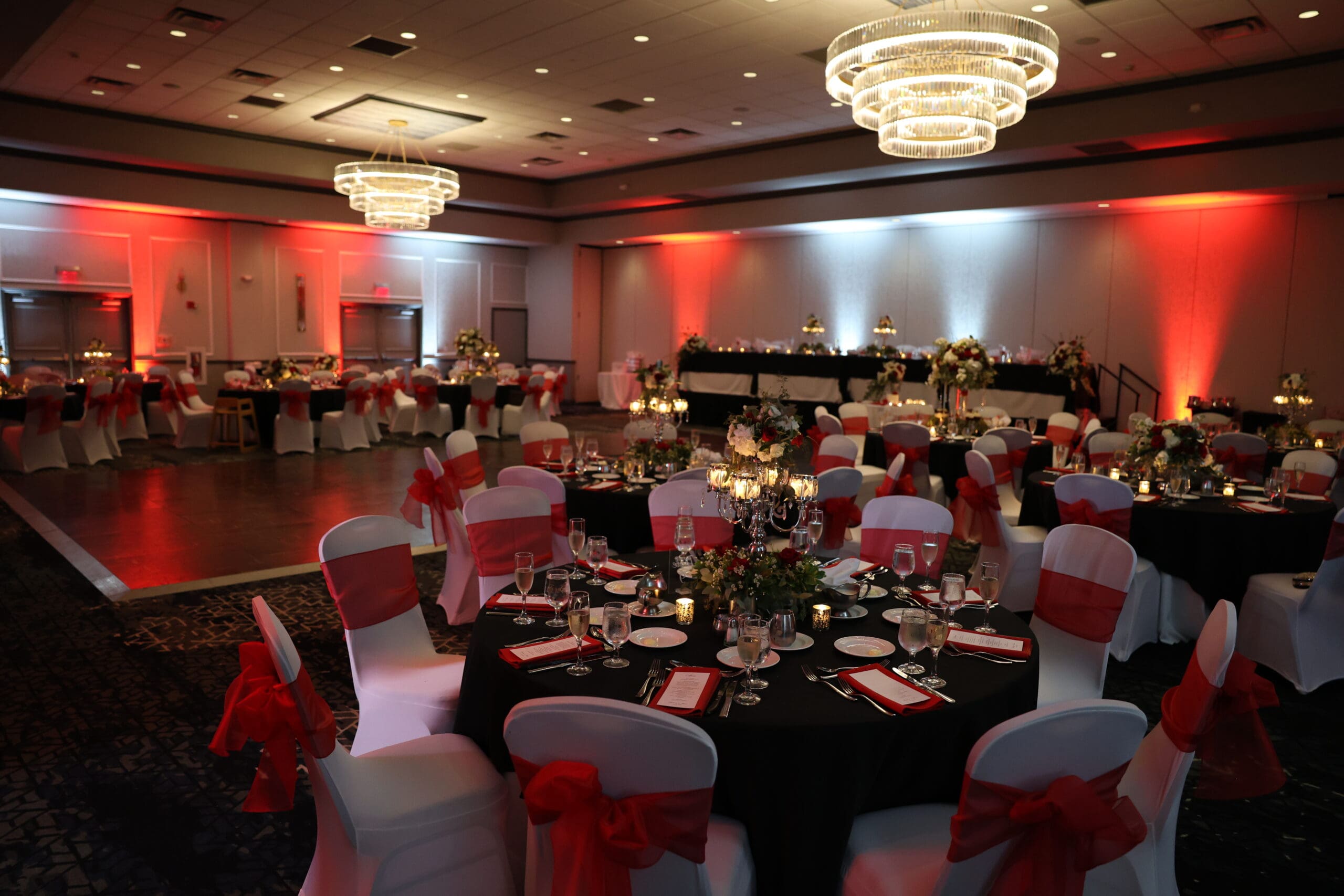 Elegant wedding reception ballroom with round tables, red chair sashes, candlelit centerpieces, and chandelier lighting.