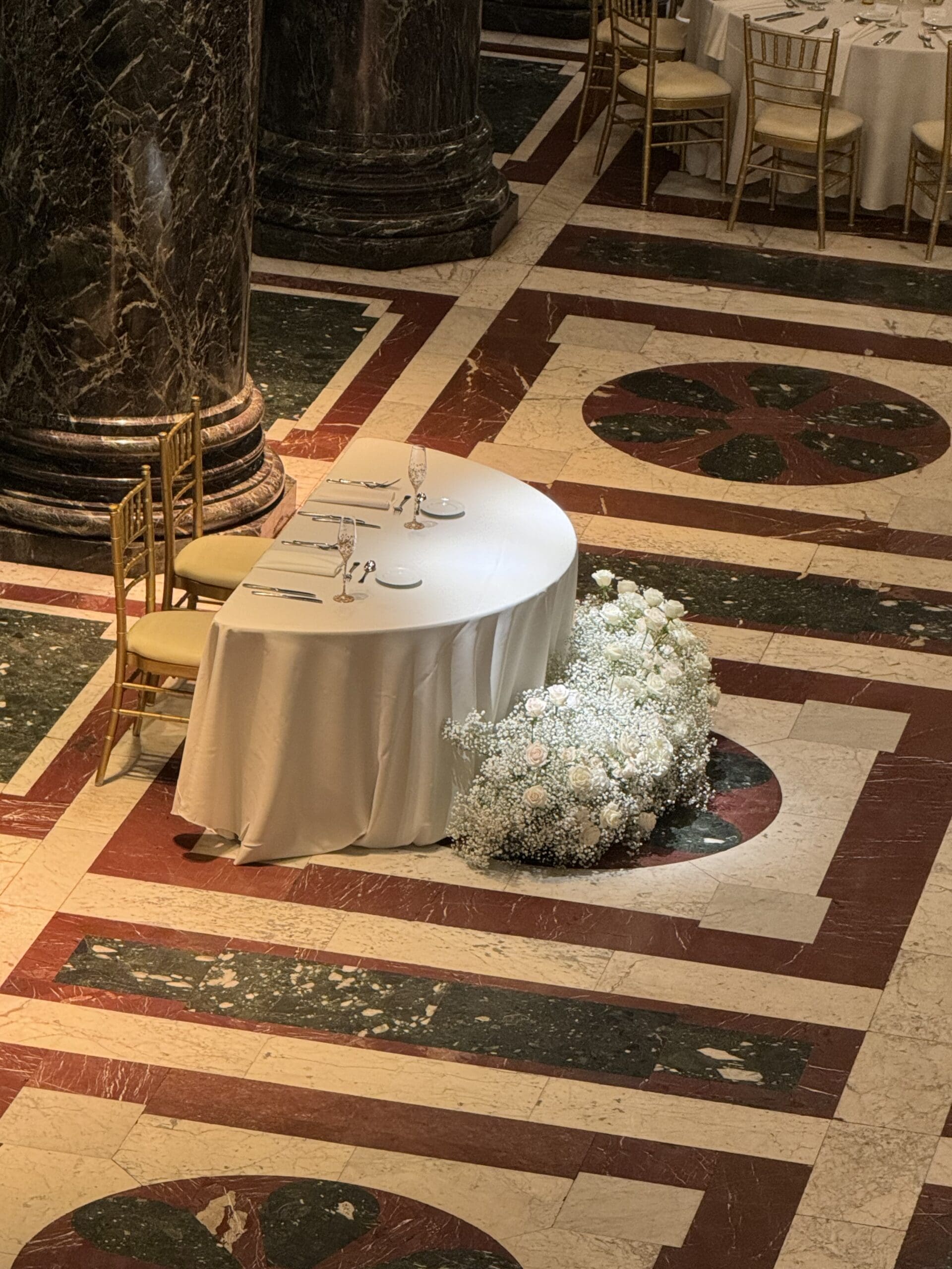 Elegant sweetheart table with white linens and floral arrangement in a grand marble ballroom.<br />
